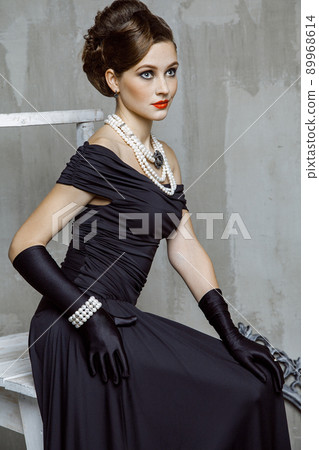 Beautiful young fashion caucasian model in black long dress and fashion makeup and beautiful hairdo posing sitting on the chair and looking at camera. Studio shot. 89968614