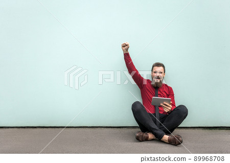 Working in outdoor, sits on the ground. Young adult bearded businessman work in tablet, hand up and shout with smile. Outdoor shot on gray wall. Working in outdoor, sits on the ground. Young adult bearded businessman work in tablet, hand up and shout with smile. Outdoor shot on gray wall. 89968708