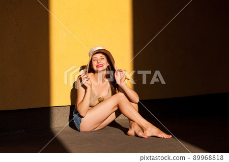 Funny young model sitting on ground, looking at camera and toothy smile. Isolated on yellow wall, outdoor 89968818