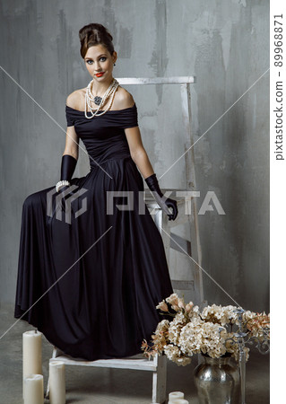 Beautiful young fashion caucasian model in black long dress and fashion makeup and beautiful hairdo posing sitting on the chair and looking at camera. Studio shot. 89968871