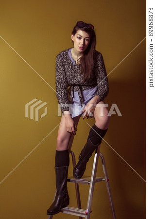 Beautiful young asian model in blue denim skirt and black blouse and fashion makeup posing sitting on stepladder and looking at camera. Studio shot. High-fashion photo. 89968873