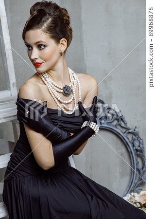 Beautiful young fashion caucasian model in black long dress and fashion makeup and beautiful hairdo posing sitting on the chair and looking at camera. Studio shot. 89968878