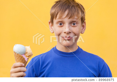 Blonde beautiful boy with freckles and blue t-shirt holds ice cream looking at camera with smile and wonder face . Studio shot, isolated on a yellow background. Blonde beautiful boy with freckles and blue t-shirt holds ice cream looking at camera with smile and wonder face . Studio shot, isolated on a yellow background. 89968975