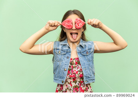 Beautiful teenager girl with blondie long hair, looking at camera and tongue out , holds two lolipop, having funny look. Isolated studio shot on light green background. Beautiful teenager girl with blondie long hair, looking at camera and tongue out , holds two lolipop, having funny look. Isolated studio shot on light green background. 89969024