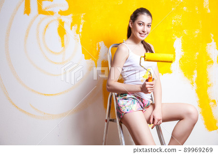 Beautiful young caucasian model in coloured shorts and white shirt and posing holding outrigger in her hands, painting the wall and looking at camera. Studio shot. Beautiful young caucasian model in coloured shorts and white shirt and posing holding outrigger in her hands, painting the wall and looking at camera. Studio shot. 89969269
