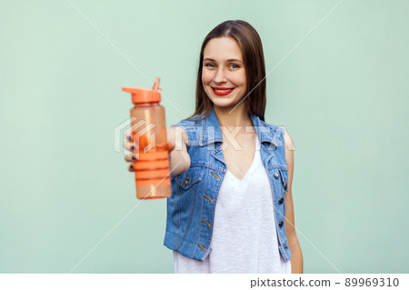 Beautiful young woman with freckles and denim jacket in casual style holding a bottle of water and looking at camera with big eyes and toothy smile. Studio shot, isolated on a green background 89969310