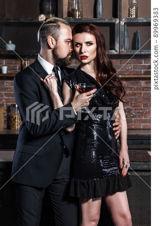 Well dressed famous couple in loft interior. Man in classic style and tie, woman in black dress. Couple holding glasses of vine. Studio shot, brick wall Well dressed famous couple in loft interior. Man in classic style and tie, woman in black dress. Couple holding glasses of vine. Studio shot, brick wall 89969383