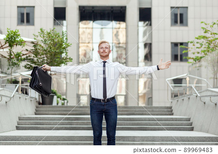 Celebrating success. Young adult red hair and bearded businessman keeping arms raised and expressing positivity while standing outdoors with office building in the background Celebrating success. Young adult red hair and bearded businessman keeping arms raised and expressing positivity while standing outdoors with office building in the background 89969483