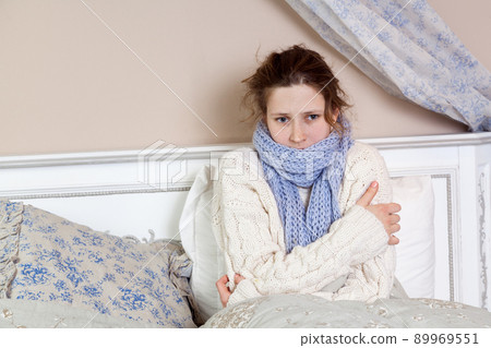 Sad alone young woman in white sweater and blue scarf feeling cold sick and resting home in bed. indoor studio shot. Sad alone young woman in white sweater and blue scarf feeling cold sick and resting home in bed. indoor studio shot. 89969551