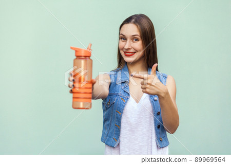 The pretty young woman with freckles in casual style holding a bottle of water and show the bottle with finger and looking at camera with toothy smile. Studio shot, isolated on a green background. 89969564