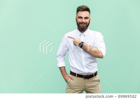 Businessman pointing copy space. Handsome young adult man with beard in white shirt looking at camera and pointing away while standing isolated on light green background Businessman pointing copy space. Handsome young adult man with beard in white shirt looking at camera and pointing away while standing isolated on light green background 89969582