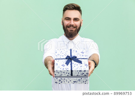 Businessman holding gift box and looking at camera and toothy smile. On light green background. Studio shot 89969753
