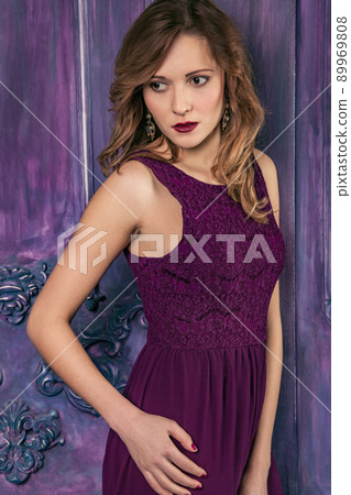 Fashion model posing in red dress near armchair..Beautiful young fashion caucasian model in violet long dress and fashion makeup and long hair posing standing near the armchair and looking at camera Fashion model posing in red dress near armchair..Beautiful young fashion caucasian model in violet long dress and fashion makeup and long hair posing standing near the armchair and looking at camera 89969808