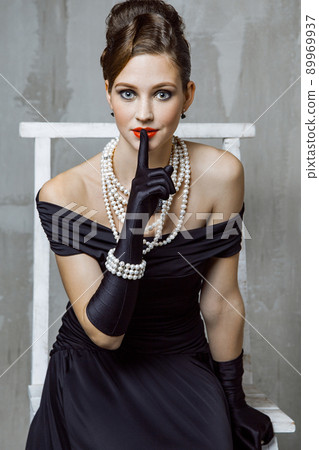 Beautiful young fashion caucasian model in black long dress and fashion makeup and beautiful hairdo posing sitting on the chair and looking at camera. Studio shot. Beautiful young fashion caucasian model in black long dress and fashion makeup and beautiful hairdo posing sitting on the chair and looking at camera. Studio shot. 89969937