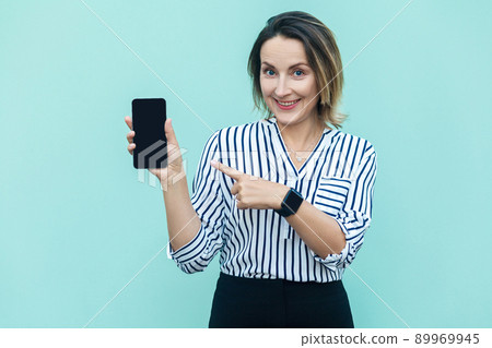 Technologies concept. Cute blonde business woman pointing finger to new smart phone and looking at camera with toothy smile. Outdoor shot, blue background. Technologies concept. Cute blonde business woman pointing finger to new smart phone and looking at camera with toothy smile. Outdoor shot, blue background. 89969945