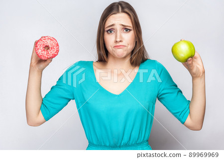 Beautiful young woman with freckles in green dress, confussed and trying to make choice between apple and donut. studio shot on light gray background 89969969