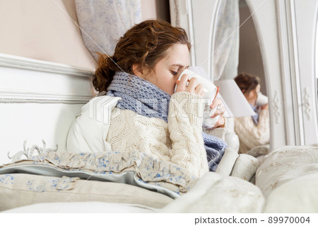 Sick woman with cup of tea. Closeup image of young frustrated woman in knitted blue scarf holding and drinking a cup of tea while sitting in bed of her room. 89970004