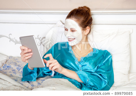 Beautiful woman with green nightie and white facial mask resting on bed and holding tablet before sleeping. healthcare and beauty treatment and technology concept. studio shot. . Beautiful woman with green nightie and white facial mask resting on bed and holding tablet before sleeping. healthcare and beauty treatment and technology concept. studio shot. . 89970085