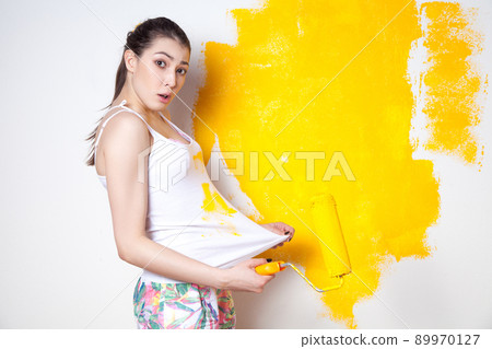 Beautiful young caucasian model in coloured shorts and white shirt and posing holding outrigger in her hands, painting the wall and looking at camera. Studio shot. 89970127