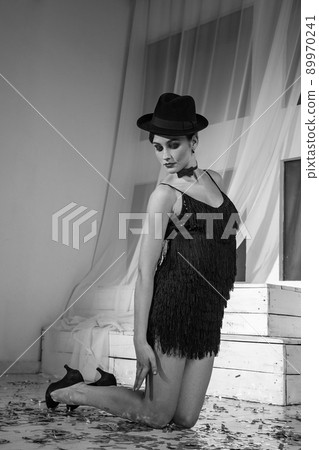 Beautiful young caucasian dancer in black dress and fashion makeup and hat posing standing on her knees and looking into the distance. Studio shot. Black and white photo. 89970241