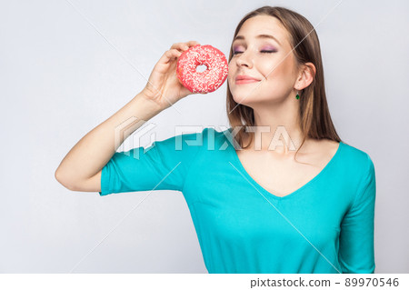 Portrait of pleasure dreaming young beautiful girl in blue blouse standing, holding and showing pink donut with closed eyes in grey background. Indoor, isolated, copy space, studio shot 89970546