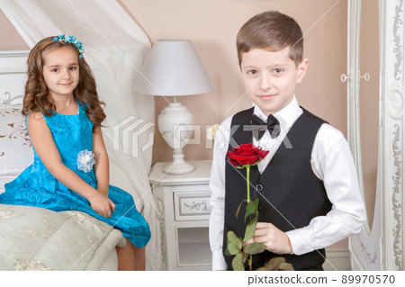 Beautiful young prince in tuxedo suit and princess holding rose in blue dress and fashion band posing sitting on the bed in bright room and looking into the distance. Love story. Studio shot. Beautiful young prince in tuxedo suit and princess holding rose in blue dress and fashion band posing sitting on the bed in bright room and looking into the distance. Love story. Studio shot. 89970570