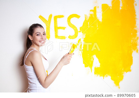 Beautiful young caucasian model in coloured shorts and white shirt and posing holding outrigger in her hands wrote Yes on wall, painting the wall and looking at camera. Studio shot. 89970605