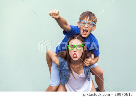 Crazy emotions and feelings. Super man style! The freckled brother climbed up on the back of a older cute sister. Making funny crazy face, tongue out and looking at camera. Indoor studio shot Crazy emotions and feelings. Super man style! The freckled brother climbed up on the back of a older cute sister. Making funny crazy face, tongue out and looking at camera. Indoor studio shot 89970613