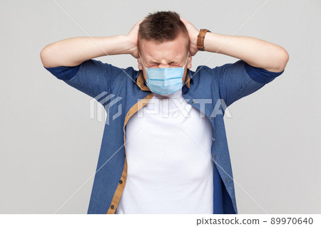 Headache. Portrait of young man in casual style with surgical medical mask standing and holding his painful head. medical and health care concept. indoor studio shot, isolated on gray background. Headache. Portrait of young man in casual style with surgical medical mask standing and holding his painful head. medical and health care concept. indoor studio shot, isolated on gray background. 89970640