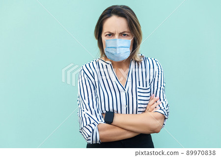 Portrait of angry middle aged woman with surgical medical mask standing with crossed arms and looking at camera frowning. medicine, health care concept. indoor studio shot isolated on blue background. 89970838