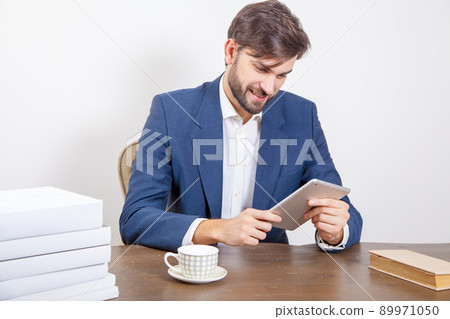 Technology, people and business concept - handsome man with beard and brown hair and blue suit and tablet pc computer and some books in the office holding tablet and working with smile. .Isolated on Technology, people and business concept - handsome man with beard and brown hair and blue suit and tablet pc computer and some books in the office holding tablet and working with smile. .Isolated on 89971050