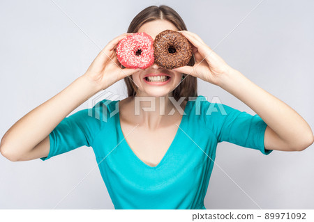 Positive young smiling girl in blue blouse standing, holding, covering eyes with pink and chocolate donuts,looking through donuts like making glasses on grey background. Indoor, isolated, copy space 89971092