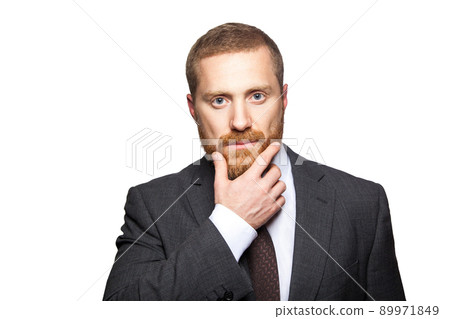 Closeup portrait of serious calm handsome businessman with facial beard in black suit standing, relaxed, touching his face and looking at camera. indoor studio shot isolated on white background. Closeup portrait of serious calm handsome businessman with facial beard in black suit standing, relaxed, touching his face and looking at camera. indoor studio shot isolated on white background. 89971849