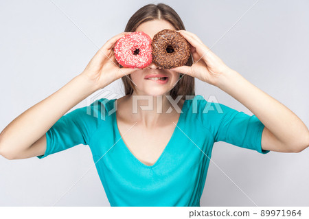 Young crazy girl bite one lips in blue blouse standing, holding, covering eyes with pink and chocolate donuts,looking through donuts like making glasses on grey background. Indoor,isolated, copy space Young crazy girl bite one lips in blue blouse standing, holding, covering eyes with pink and chocolate donuts,looking through donuts like making glasses on grey background. Indoor,isolated, copy space 89971964