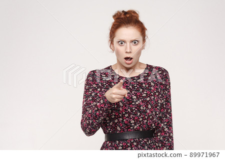 Redhead woman wearing flowers dress, opening mouths widely, having surprised shocked looks, pointing finger at camera. Isolated studio shot on gray background Redhead woman wearing flowers dress, opening mouths widely, having surprised shocked looks, pointing finger at camera. Isolated studio shot on gray background 89971967