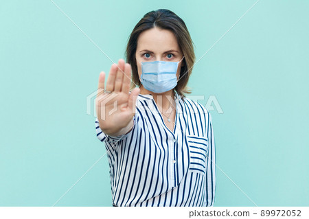 Stop, don't come any closer. Portrait of serious middle aged woman with surgical medical mask standing and showing stop sign. medicine, health care concept. indoor shot isolated on blue background. Stop, don't come any closer. Portrait of serious middle aged woman with surgical medical mask standing and showing stop sign. medicine, health care concept. indoor shot isolated on blue background. 89972052