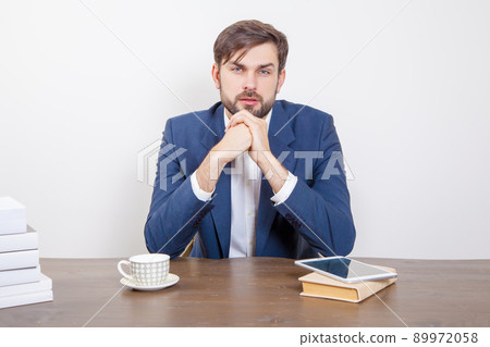 Technology, people and business concept - handsome man with beard and brown hair and blue suit and tablet pc computer and some books sitting in the office looking at camera with serious face Technology, people and business concept - handsome man with beard and brown hair and blue suit and tablet pc computer and some books sitting in the office looking at camera with serious face 89972058