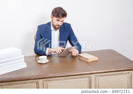 Technology, people and business concept - handsome man with beard and brown hair and blue suit and tablet pc computer and some books holding tablet, working and sitting in the office. .Isolated on Technology, people and business concept - handsome man with beard and brown hair and blue suit and tablet pc computer and some books holding tablet, working and sitting in the office. .Isolated on 89972151