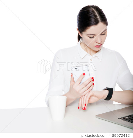 Portrait of attractive brunette businesswoman in white shirt sitting, holding smartphone mobile and looking at display of her smartwatch and reading something. studio shot isolated in white background 89972412