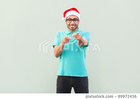 Hey you! Its christmas man! Bearded man in santa hat and glasses, pointing finger and looking at camera with toothy smile. On gray background. Indoor, studio shot Hey you! Its christmas man! Bearded man in santa hat and glasses, pointing finger and looking at camera with toothy smile. On gray background. Indoor, studio shot 89972456