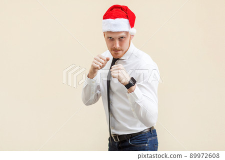 Boxing. Young adult ginger businessman on red santa hat, ready for fight on light orange background. Studio shot 89972608