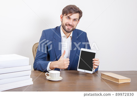 Technology, people and business concept - handsome man with beard and brown hair and blue suit and tablet pc computer and some books in the office, holding tablet and showing display screen with thumb Technology, people and business concept - handsome man with beard and brown hair and blue suit and tablet pc computer and some books in the office, holding tablet and showing display screen with thumb 89972830