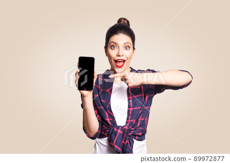 Young beautiful shocked woman in casual style holding phone looking at camera and showing phone display. studio shot on beige background. 89972877
