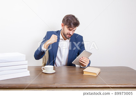 Technology, people and business concept - handsome man with beard and brown hair and blue suit and tablet pc computer and some books sitting in the office angry and unhappy. .Isolated on white 89973005