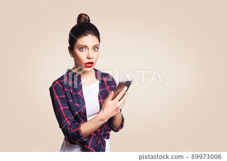 Young beautiful woman in casual style holding phone and surprised. shocked and looking at camera. studio shot on beige background. Young beautiful woman in casual style holding phone and surprised. shocked and looking at camera. studio shot on beige background. 89973006