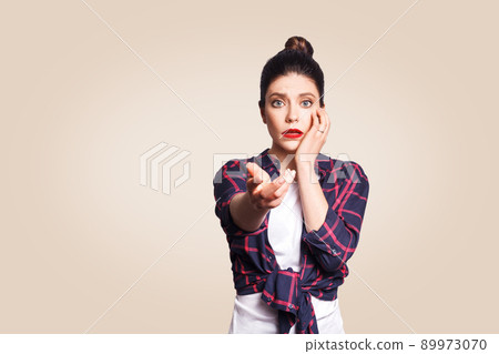 Scared girl with bun hair knot having scared and frightened look, keeping mouth open, touching face and pointing hand at camera. 89973070