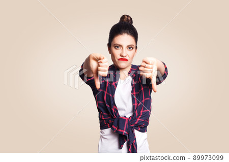 Dislike. Young unhappy upset girl with casual style and bun hair thumbs down her finger, on beige blank wall with copy space looking at camera with toothy smile. focus on face. 89973099