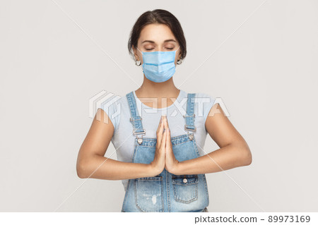 relaxation and yoga meditation. Portrait of calm young brunette woman with surgical medical mask in denim overalls standing in yoga pose and meditating. indoor studio shot isolated on gray background. relaxation and yoga meditation. Portrait of calm young brunette woman with surgical medical mask in denim overalls standing in yoga pose and meditating. indoor studio shot isolated on gray background. 89973169