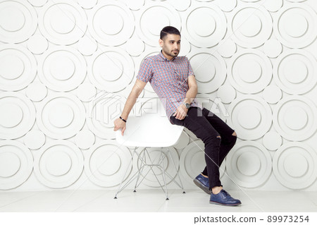 Young bearded fashion model in casual style is posing near white circle wall background. studio shot. sitting on chair and looking at away. 89973254