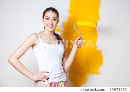 Beautiful young caucasian model in coloured shorts and white shirt and posing holding outrigger in her hands, painting the wall and looking at camera. Studio shot. 89973283